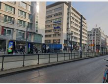 Straßenansicht einer modernen städtischen Umgebung mit mehreren mehrstöckigen Gebäuden, darunter ein Starbucks und Geschäfte, sowie einer Straßenbahnhaltestelle mit Fahrgästen und Werbedisplays. Im Vordergrund führen Straßenbahngleise entlang eines Bürgersteigs, daneben eine mehrspurige Straße.