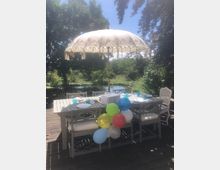 Ein gedeckter Gartentisch auf einer Holzterrasse mit weißen Stühlen und einem dekorativen Sonnenschirm mit Muster. Im Hintergrund ist ein Teich und grüne Vegetation zu sehen; Luftballons schmücken einen der Stühle.