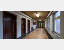 Ein schmaler, länglicher Flur mit braunem Linoleumboden, braunen Türen auf der linken Seite und großen rechteckigen Fenstern auf der rechten Seite, die Tageslicht hereinlassen. Am Ende des Flurs befinden sich doppelte Glastüren; die Decke ist beige und mit rechteckigen Deckenlampen ausgestattet.