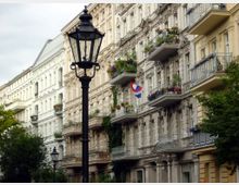 Reihe historischer Wohnhäuser mit verzierten Fassaden und Balkonen, geschmückt mit Blumen. Im Vordergrund steht eine traditionelle schwarze Straßenlaterne, umgeben von Bäumen.