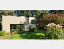 Modernes Wohnhaus mit einem flachen, rechteckigen Design, großen Glasfenstern und einer Fassade in heller Farbe. Umgeben von einem gepflegten Garten mit Rasen, Ziergräsern und dichtem Baumbestand im Hintergrund.