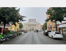 Straßenansicht mit der Volksbühne in Berlin im Hintergrund, einem großen historischen Gebäude mit Säulenfassade und Schriftzug „Volksbühne“. Im Vordergrund sind parkende Autos, Leihfahrräder und Fußgänger auf einer belebten Straße zu sehen, flankiert von Bäumen und Gebäuden.