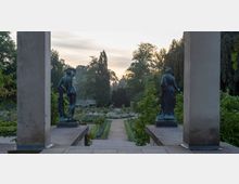Blick durch zwei steinerne Säulen auf einen Garten mit einem symmetrischen Weg, gesäumt von Blumenbeeten und Bäumen. Im Vordergrund stehen zwei bronzene Statuen auf Podesten, die jeweils eine nackte und eine bekleidete Figur darstellen.
