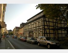 Straßenszene mit Fachwerkhaus auf der rechten Seite, flankiert von weiteren Gebäuden in verschiedenen Baustilen. Geparkte Autos säumen die Straße, während Bäume und ein Straßenlaterne den Gehweg rechts säumen.