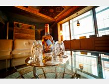 Eine moderne Lounge mit Holzwänden und großen Fenstern, die viel Tageslicht hereinlassen. Im Vordergrund steht eine Glasplatte mit einem Tablett, darauf eine Whiskeyflasche der Marke "Stork Club" und zwei Gläser.