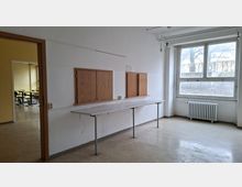 Leerer Raum in einem älteren Schul- oder Bürogebäude mit weiß gestrichenen Wänden, großem Fenster und Heizkörper darunter. An der Wand befindet sich eine lange Ablagefläche mit zwei Holzklappen-Fensternischen; links führt eine offene Tür in einen weiteren Raum mit Tischen.