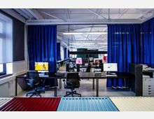 Moderner Büroarbeitsplatz in einem offenen Raum mit sichtbaren weißen Stahlträgern an der Decke, großen Tischen mit Monitoren und Laptops, sowie blauen Vorhängen, die Bereiche voneinander abtrennen. Fenster auf der linken Seite lassen Tageslicht herein, während der Raum in einem minimalistischen Design gestaltet ist.