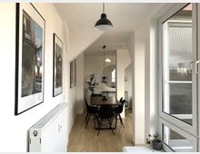 Heller, moderner Essbereich in einer Wohnung mit einem langen Holztisch und schwarzen Stühlen. An der linken Wand sind gerahmte Bilder, rechts sorgen große Fenster für viel Tageslicht; im Hintergrund sind Pflanzen und eine runde Wandspiegel-Dekoration sichtbar.