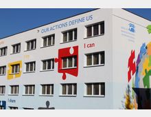Außenansicht eines modernen Schulgebäudes mit farbenfrohen Wandgestaltungen, darunter große Puzzleteile und motivierende Schriftzüge wie "I can" und "Our actions define us"; rechts oben befindet sich das Logo der Berlin Brandenburg International School.