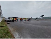 Ein kleiner Flugplatz mit einem geparkten weißen Privatjet auf dem Rollfeld. Im Vordergrund arbeiten Personen in Warnwesten an einer Film- oder Fotoausrüstung neben einem Transportfahrzeug, während links eine Gebäudewand und rechts Hangars zu sehen sind.