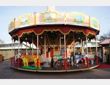 Historisches Karussell mit bemalten Holzpferden und farbenfrohen Dekorationen, umgeben von roten und goldenen Stoffbannern; im Hintergrund sind moderne Gebäude und ein Weihnachtsmarktstand zu sehen.