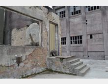 Ruine eines Industriegebäudes mit freiliegenden Ziegelwänden, zerbrochenen Fensterscheiben und bröckelndem Putz. Im Vordergrund befindet sich eine kleine Betontreppe und ein offener Türrahmen, dahinter ist eine größere Fassade mit mehrfachen Fensteröffnungen sichtbar.
