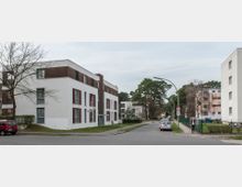 Straßenszene in einem Wohngebiet mit modernen, mehrstöckigen Wohngebäuden aus weißem Putz und Holzfassaden-Elementen, umgeben von Grünflächen und hohen Bäumen. Am Straßenrand stehen geparkte Autos, daneben Gehwege mit vereinzelten Fußgängern.