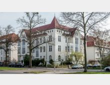 Eckgebäude eines Wohnhauses im klassischen Stil mit weißer Fassade und rotem Satteldach, umgeben von kahlen Bäumen und parkenden Autos entlang einer ruhigen Straßenkreuzung.
