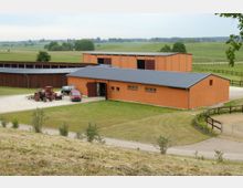Weitläufiger Bauernhof mit einem orangefarbenen Gebäude mit Flachdach im Vordergrund, neben einem Hof mit einem Traktor und einem Anhänger. Im Hintergrund befinden sich weitere landwirtschaftliche Gebäude und grüne Felder, umgeben von Zäunen und Bäumen.