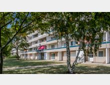 Wohnhaus mit mehreren Etagen und Balkonen, umgeben von Bäumen und einer Rasenfläche. Im Vordergrund sind Birken und ein gepflasterter Gehweg zu sehen, im Hintergrund ragt ein pinker Sonnenschirm auf einem der Balkone hervor.