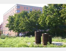 Grüne Parkanlage mit dichten Bäumen und einer Wiese im Vordergrund, auf der zylindrische, brunnenartige Skulpturen stehen. Im Hintergrund ist ein mehrstöckiges, rosafarbenes Wohngebäude zu sehen.