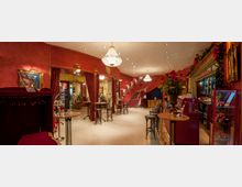 Eleganter Innenraum eines Theaters oder Varietés mit roten Wänden, Kronleuchtern und goldenen Akzenten. Ein geschwungener Treppenaufgang, Barhocker an einer Theke und dekorative Elemente wie Vorhänge und Blumenschmuck prägen das Ambiente.