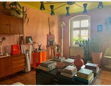 Ein heller Raum mit rosa Wänden und gelber Decke, eingerichtet als kreativer Arbeitsbereich. Zu sehen sind ein Tisch mit Büchern und Dekorationen, eine Staffelei mit einem Gemälde, zahlreiche Pflanzen sowie ein Holzschrank und eine orangefarbene Rollkommode.