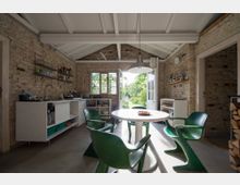 Innenraum einer rustikalen Küche mit freiliegenden Backsteinwänden und einem hohen, weißen Holzgebälk. Ein runder Esstisch mit grünen Stühlen steht im Raum, der durch eine geöffnete Terrassentür Blick auf einen sonnigen Garten bietet.