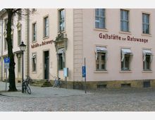 Eine Straßenansicht eines historischen Gebäudes mit der Aufschrift „Gaststätte zur Ratswaage“. Das helle Gebäude hat hohe Fenster mit weißen Markisen, eine steinerne Eingangstreppe und ein dekoratives Türportal; davor stehen Fahrräder und Laternen auf dem gepflasterten Gehweg.