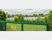 Grünes Metallgittertor mit einem gelben Schild, auf dem "Ausfahrt freihalten" steht, im Vordergrund; dahinter eine Rasenfläche, hohe Büsche und Schilf am Ufer eines Sees, umgeben von Bäumen unter einem wolkigen Himmel.