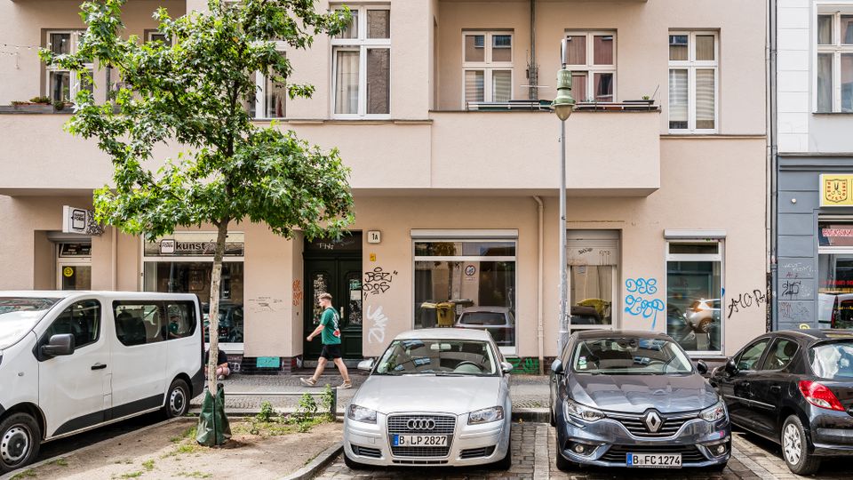 Straßenansicht eines hell verputzten Mehrfamilienhauses mit mehreren Fenstern und Balkonen sowie Ladenfronten im Erdgeschoss. Davor stehen ein Straßenbaum, eine Laterne und mehrere parkende Autos entlang des Bordsteins.