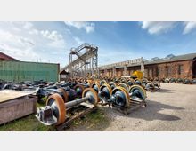 Ein Außenbereich eines Industriegeländes mit zahlreichen gelagerten Eisenbahnradsätzen auf Holzpaletten, einem grünen Container, einem Förderbandgerüst und einem Backsteingebäude im Hintergrund. Der Himmel ist blau mit einigen Wolken.