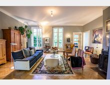 Wohnzimmer mit Holzboden, einem blauen Sofa, verschiedenen Sesseln und einem weißen Couchtisch auf einem gemusterten Teppich. Große Fenster mit Blick in einen Garten, helle Wände, antike Holzschränke und dekorative Elemente runden den Raum ab.