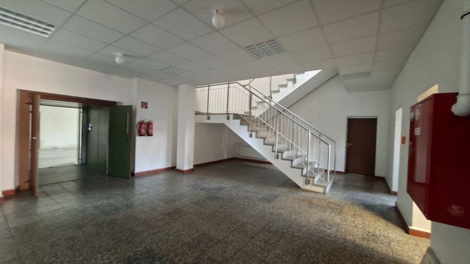 Geräumiges Treppenhaus in einem Gebäude mit gefliester Bodenfläche, weißer Rasterdecke und einer Treppe mit Metallgeländer. Links befindet sich ein offener Durchgang zu einem weiteren Raum, rechts sind eine Tür sowie ein roter Feuerlöschkasten und Feuerlöscher an der Wand zu sehen.