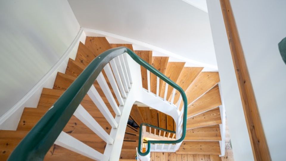 Blick von oben in ein helles Treppenhaus mit gewendelten Holzstufen und weißen Geländern. Ein grüner Handlauf führt entlang der Treppe, umgeben von weißen Wänden und Holzdetails.