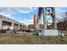 Industriebrache mit überwuchertem Gelände, alten Metallkonstruktionen und Backsteingebäuden, darunter ein runder Backsteinturm im Hintergrund. Der Himmel ist blau mit einigen Wolken und umgibt die verlassene Szenerie.