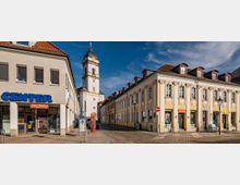 Straßenszene mit einem historischen gelben Gebäude rechts, verziert mit weißen Stuckelementen und kleinen Geschäften im Erdgeschoss. Im Hintergrund ist ein weißer Kirchturm mit grüner Kuppel zu sehen; links steht ein modernes Gebäude mit einem Laden mit der Beschriftung "Center".