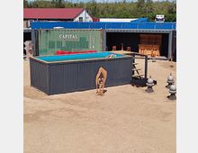 Ein rechteckiger Pool in einem umgebauten Schiffscontainer steht auf sandigem Boden, umgeben von weiteren Schiffscontainern sowie hölzernen und metallenen Dekorationselementen. Im Hintergrund sind Bäume, sowie ein Gebäude mit rotem Dach sichtbar.