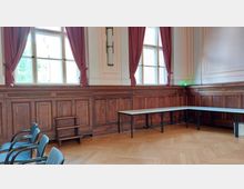 Ein historischer Raum mit Holztäfelung an den Wänden, großen Fenstern mit roten Vorhängen und einem Fischgrät-Holzboden. Der Raum ist mit einfachen Tischen, Stühlen und einer kleinen Holztreppe ausgestattet.