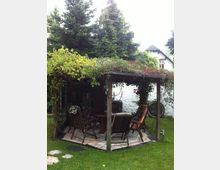 Ein mit Kletterpflanzen bewachsener Pavillon in einem Garten mit einer Sitzgruppe aus Holzmöbeln auf einem gefliesten Boden; im Hintergrund sind Bäume und ein weißes Haus mit Schrägdach zu sehen.