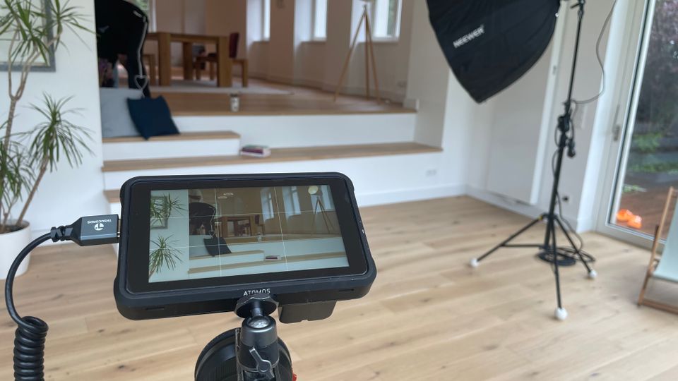 Heller Innenraum mit hellen Holzböden und großen Fenstern; im Hintergrund liegt eine erhöhte Sitz- oder Podestzone mit Tisch und Stehlampe. Im Vordergrund stehen Film-/Fotogeräte wie ein Kameramonitor auf Stativ und eine große Softbox-Leuchte neben einer Glastür zum Garten.