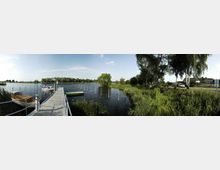 Panoramaaufnahme eines kleinen Hafens an einem See mit schmalem Steg, an dem mehrere kleine Boote festgemacht sind. Die Umgebung besteht aus dichter Vegetation, Bäumen und Gräsern; im Hintergrund sind vereinzelte Gebäude und ein parkendes Fahrzeug sichtbar.