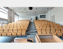 Ein moderner Hörsaal mit mehreren Reihen hellbrauner, klappbarer Sitze, die stufenförmig angeordnet sind. Im Vordergrund befindet sich ein Rednerpult mit Mikrofon, während an der Rückwand des Raums ein Fenster in einem Technikraum erkennbar ist.
