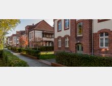 Reihen von Backstein-Mehrfamilienhäusern mit weißen Fassadenelementen und roten Dächern, flankiert von gepflegten Hecken und Gehwegen. Vereinzelte Bäume säumen die Straße, die in einer ruhigen Wohngegend liegt.