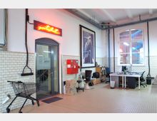 Bürobereich in einem industriellen Gebäude mit weiß gefliesten Wänden und hohen Decken. Zu sehen sind ein Schreibtisch mit Bürostühlen, ein rotes Wandpaneel mit Feuerlöscher, ein Einkaufswagen und eine Glastür, über der ein leuchtendes Neon-Schild angebracht ist.