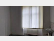 Leerer Raum mit weißen Wänden, einem großen Fenster mit vertikalen Lamellenjalousien und einem Heizkörper darunter; rechts neben dem Fenster ein kleines Waschbecken mit Kacheln an der Wand.