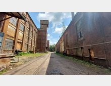 Industriegelände mit alten Backsteingebäuden, die große Fenster mit Glasbausteinen aufweisen. Eine schmale gepflasterte Straße verläuft zwischen den Gebäuden, die von Vegetation und Resten von Schienen gesäumt ist; im Hintergrund steht ein Turm.