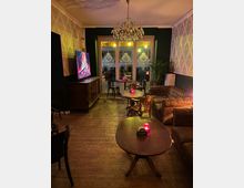 Ein gemütlicher, beleuchteter Innenraum mit Holzfußboden, eleganten Tapeten in einem floralen Muster, einem Kronleuchter und großen Fenstern mit verzierten Milchglas-Elementen. Der Raum ist möbliert mit runden und ovalen Holztischen, Vintage-Stühlen und einem Chesterfield-Sofa; kleine rote Teelichter stehen auf den Tischen.