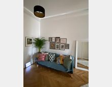 Wohnzimmer mit heller Wandfarbe. Ein Sofa mit bunter Kissenvielfalt steht vor einer Galerie aus gerahmten Bildern, daneben eine Zimmerpflanze. Ein großer Spiegel lehnt an der Wand, und ein moderner Lampenschirm hängt von der Decke. Der Boden ist mit Fischgrätparkett ausgelegt.