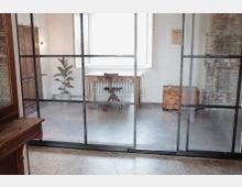 Raum mit industrieller Glaswand und minimalistischer Einrichtung. Dahinter sind ein Holztisch mit passendem Stuhl, eine Pflanze, eine Holzkiste und eine Mauer aus Ziegelsteinen sichtbar; ein Fenster sorgt für natürliches Licht.