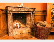 Ein gemauerter Kamin mit Marmorumrandung ist in einer gemütlichen Holzvertäfelung eingebaut. Daneben steht ein großer Weidenkorb mit Holzscheiten, und im Kamin brennt ein kleines Feuer.