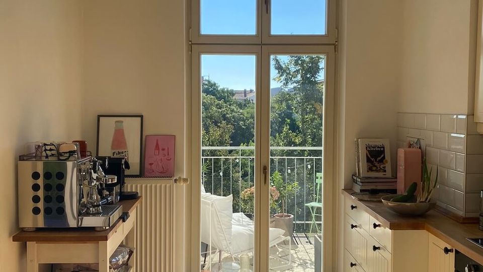 Bright, narrow kitchen with white cabinets, wooden countertops, and a small side cart, centered on tall French doors with an arched transom window. The doors open to a small balcony with railing and seating, overlooking leafy trees and nearby buildings.