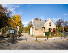 Kleines Backsteingebäude mit Spitzdach und gotischen Fensterelementen, umgeben von herbstlichen Bäumen. Links ein gepflasterter Weg mit einer Informationstafel, rechts eine Kopfsteinpflasterstraße, im Hintergrund ein Parkeingang.