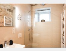 Modernes Badezimmer mit beige gefliesten Wänden und einer bodengleichen Glasdusche mit Regenduschkopf. Im Raum sind freiliegende Ziegelsteine, ein beleuchteter Spiegel, ein Heizkörper und ein kleines Fenster mit einer Pflanze sichtbar.